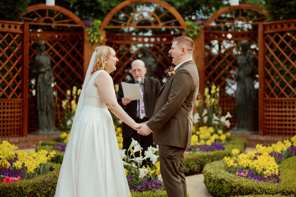 A heterosexual couple holds hands and laughs during their wedding ceremony in a garden.